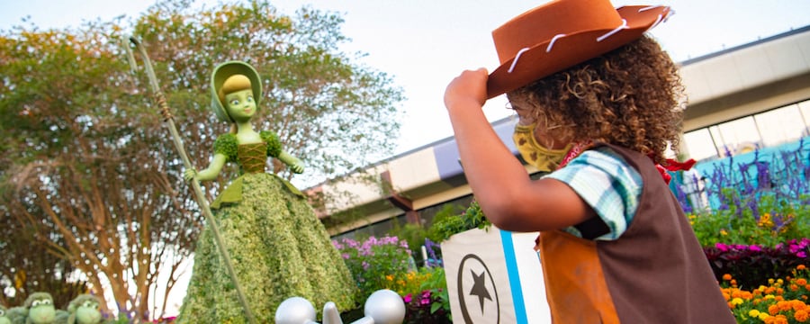 A little girl wearing a costume inspired by Jessie from Toy Story, tipping her hat at a topiary of Bo Peep