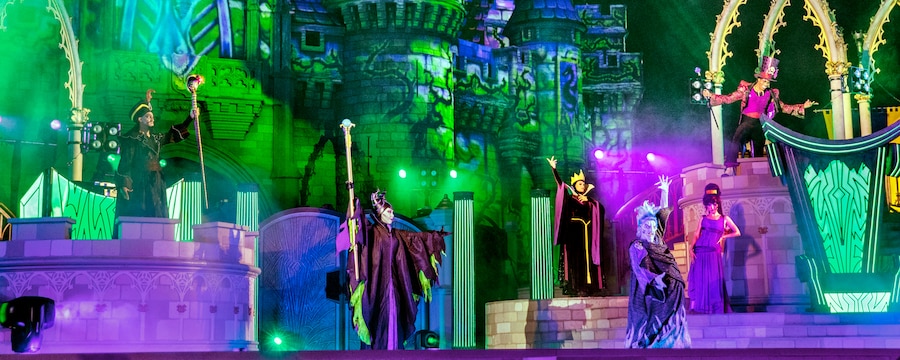 Maleficent, Hades, Meg, The Queen, Jafar and Doctor Facilier pose on stage in front of Cinderella Castle
