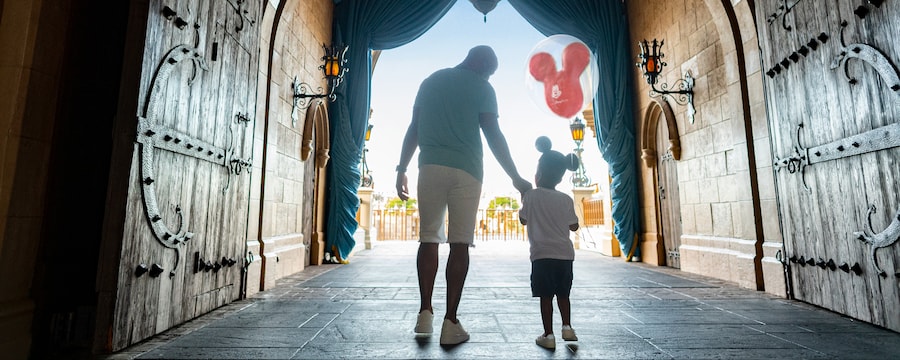 Dad and son walk through Cinderella's Castle holding a Mickey Balloon