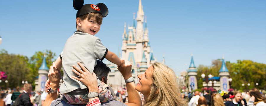 A boy wearing Mickey ears sits on top of his father’s shoulders in front of Cinderella Castle