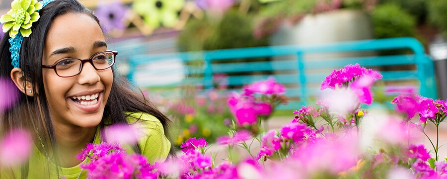 A smiling girl wearing glasses and a floral headband sits within a garden exhibit