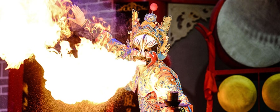 A performer in costume and mask breathes fire during a performance