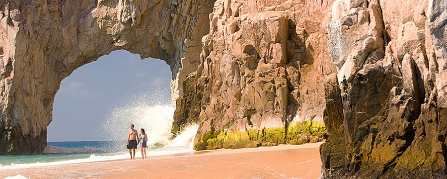 A couple holds hands while walking on the beach by El Arco in Cabo San Lucas, Mexico