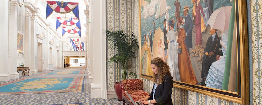 A woman types on a computer while seated on a bench beneath a large painting in a long, carpeted hallway