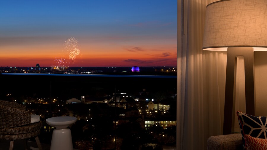 A hotel room window affords a view of fireworks exploding beyond a balcony at sunset