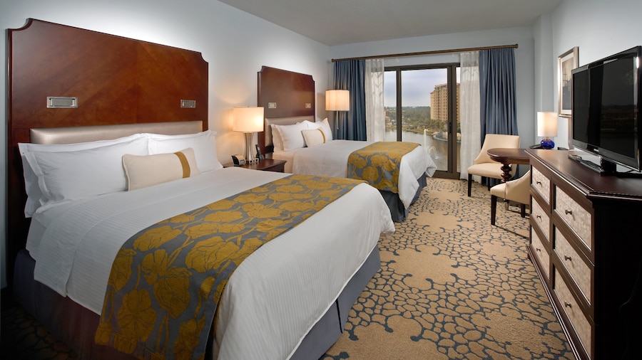 Una habitación de hotel con 2 camas Queen Size, una cómoda, sillas, una mesa y una ventana de longitud completa con vista al lago