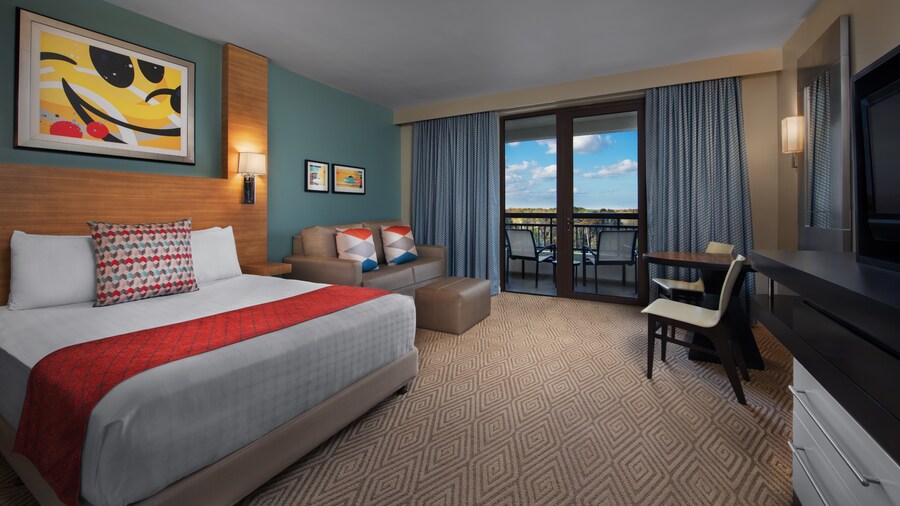 A room with a bed, 3 paintings, a couch, pillows, a TV, a table, 2 chairs and an ottoman near a balcony