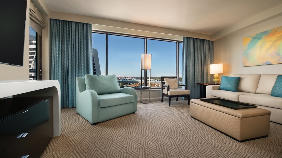 A room with a loveseat, a couch, an ottoman, a TV, drawers, a painting, a chair, 2 lamps and a window