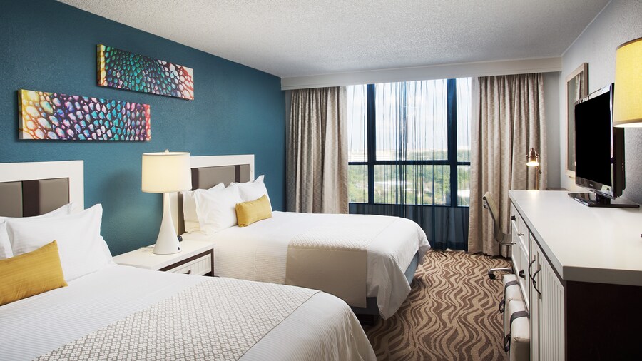 A room with 2 beds, 3 lamps, 2 paintings, a mirror, a TV, a chair and curtains