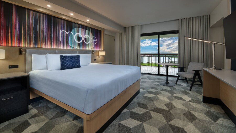 View from entryway of a king bed opposite a media console with a flat-screen TV and table and chair adjacent to a balcony