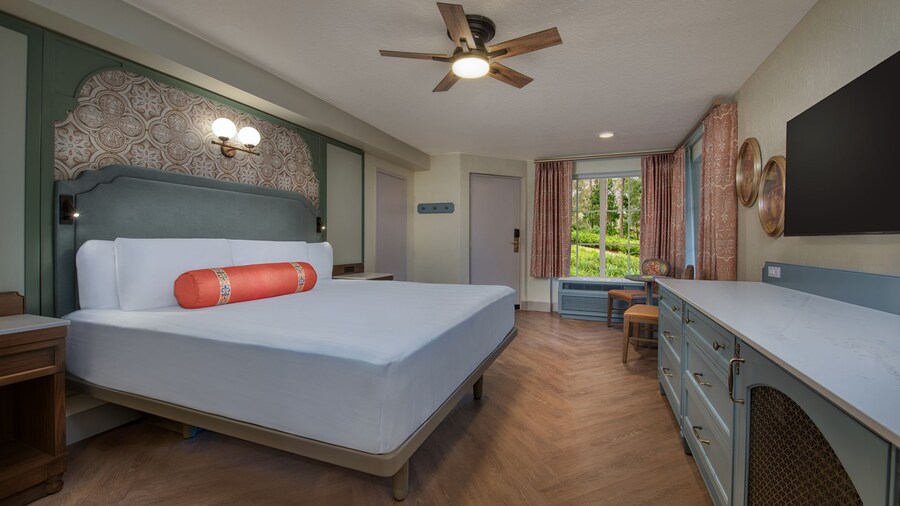 A Disney’s Port Orleans Resort hotel bedroom with a dresser, TV, ceiling fan, bed, 2 nightstands, 2 chairs and a table