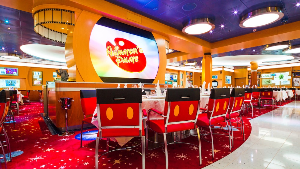 A photo of the Animator's Palate restaurant interior on a Disney Cruise Line ship. The photo features black and white walls with splashes of color, tables with chairs, and large screens displaying classic Disney animation. Guests in the foreground may be interacting with the scene or looking surprised by the transformation. (Photo Courtesy Disney Cruise Line)