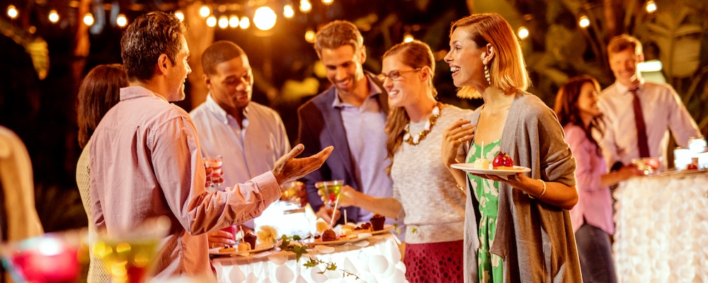 Smiling people standing around high tables draped with linens featuring dessert plates and beverages in an outdoor setting Smiling people standing around high tables draped with linens featuring dessert plates and beverages in an outdoor setting