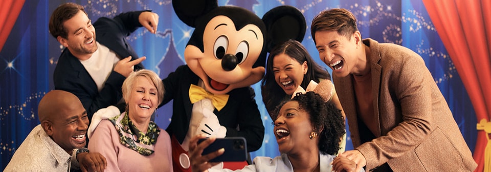 Six people are gathered around Mickey Mouse and laugh uproariously as they take a selfie
