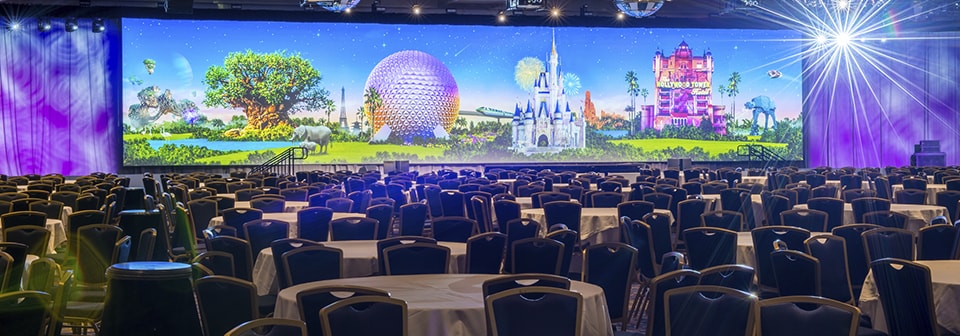 A ballroom filled with round tables and chairs, colorful lighting and monitors