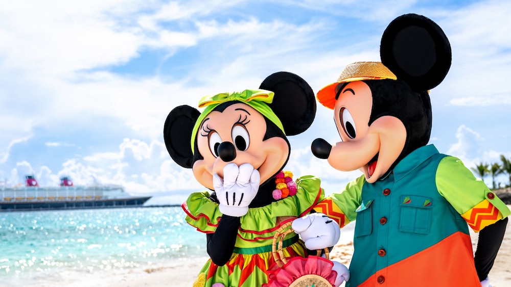 Mickey Mouse and Minnie Mouse in Junkanoo outfits standing on a Bahamian beach with a Disney Cruise Line ship on the horizon