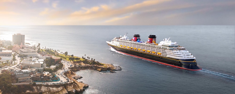 A Disney Cruise Line ship sailing past a rocky shoreline with buildings and palm trees