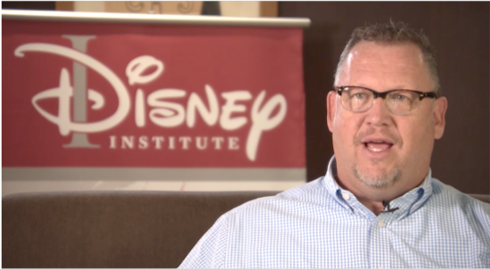A talking man seated on a couch in front of the Disney Institute sign