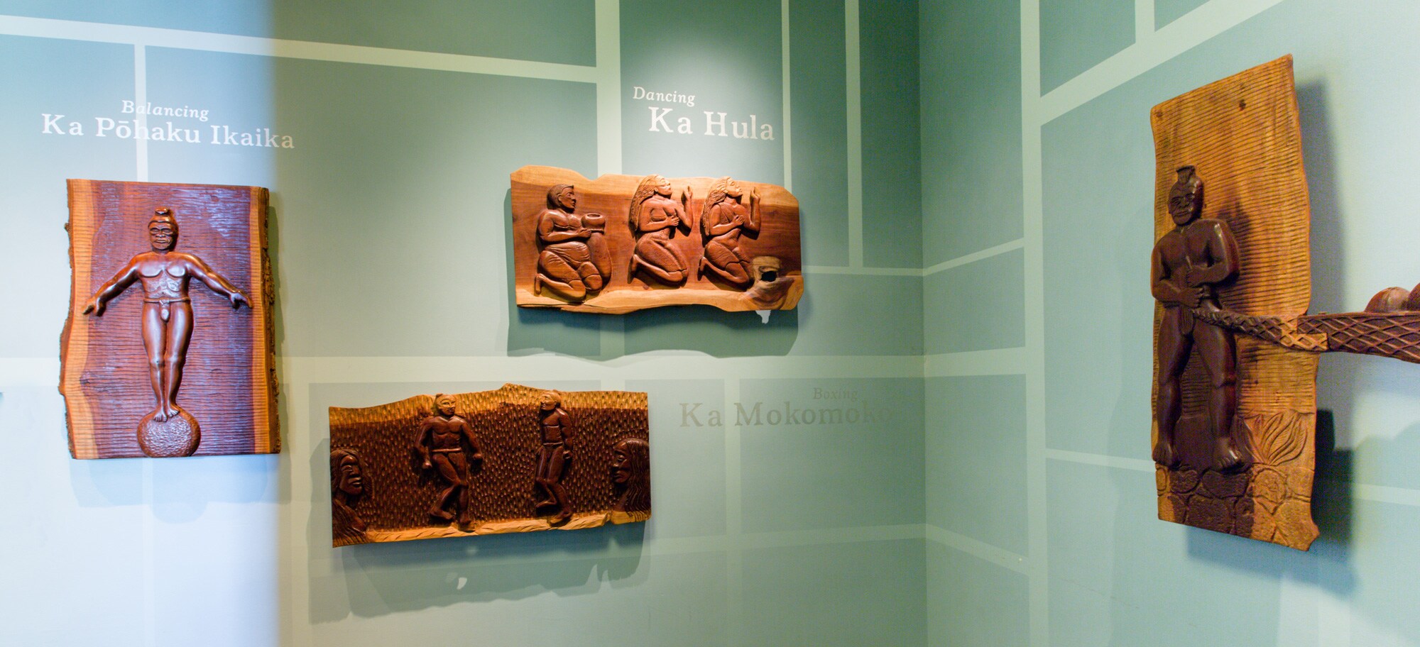 Wood relief carvings of natives engaged in various activities, with descriptive Hawaiian phrases and their English translations