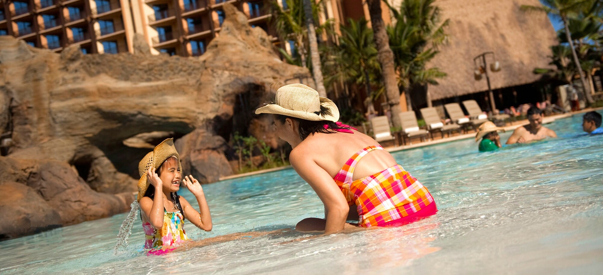 A mother and daughter, both wearing straw hats, play in the shallow end of a pool with rock caves