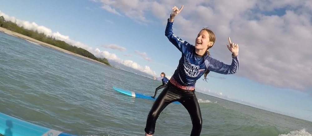 A teenage girl rides a surfboard in the ocean and gestures gleefully