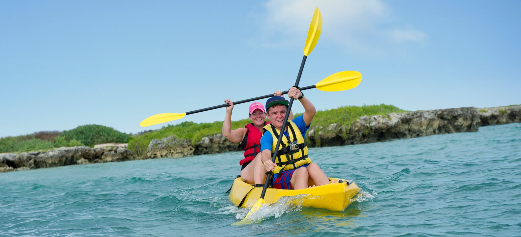 A couple paddles a kayak through the water with a rocky shore in the background