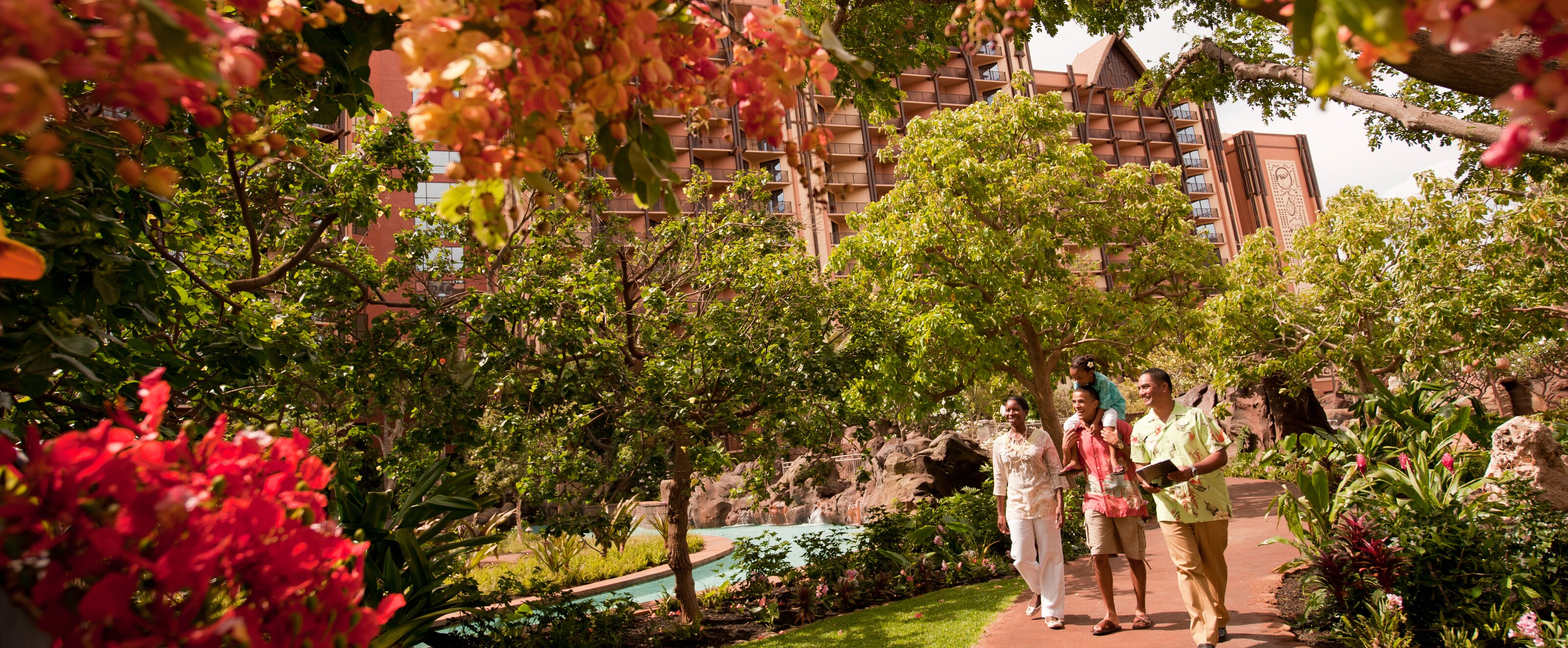 A Cast Member guides a mother and father with his young daughter sitting on his shoulders down a pathway surrounded by trees and flowers