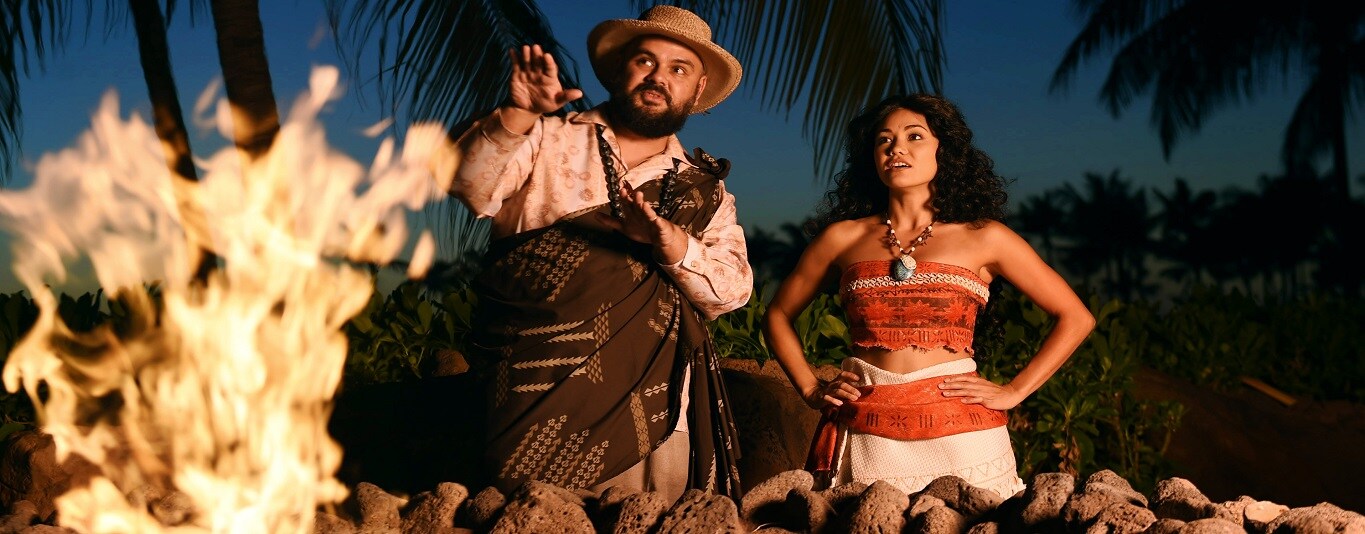 A man in a hat and sarong gestures dramatically toward a blazing fire pit while Moana watches next to him