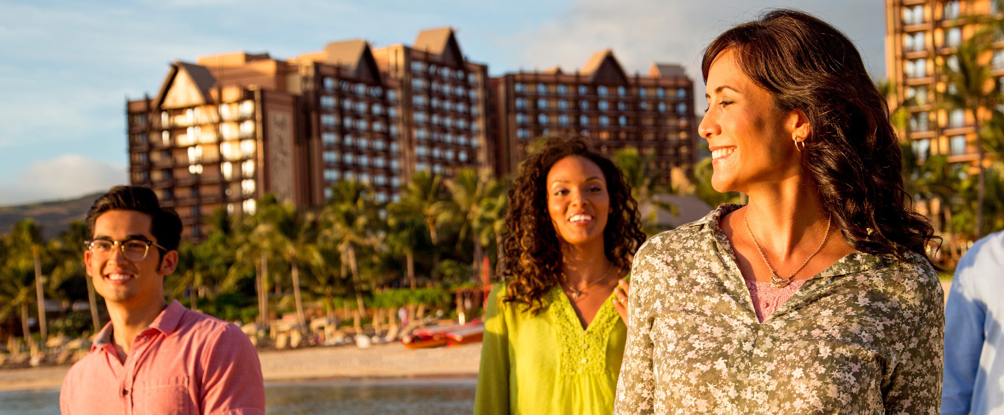 A young man and two young women walk along the beach in front of the Aulani Resort.