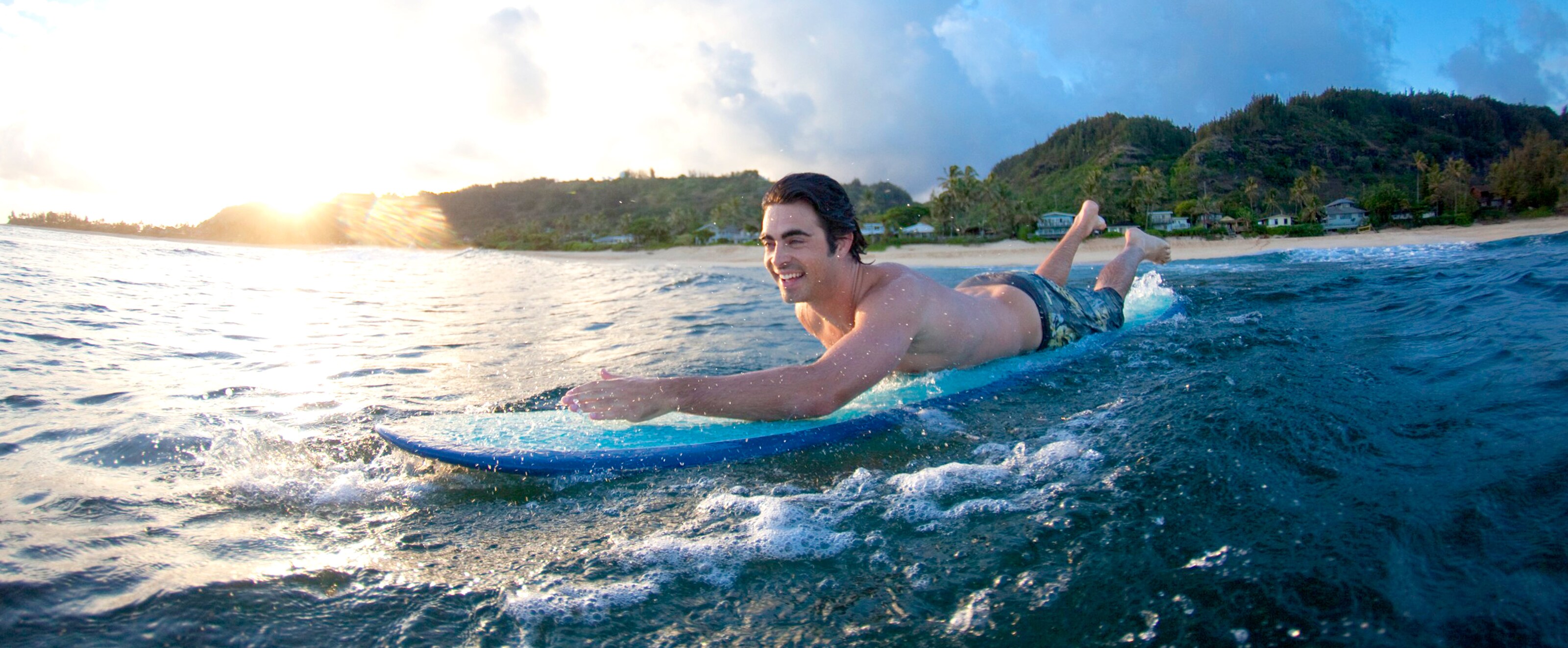A young man on a surfboard paddles out into the ocean as a coastline with tree-covered hills stretches behind him