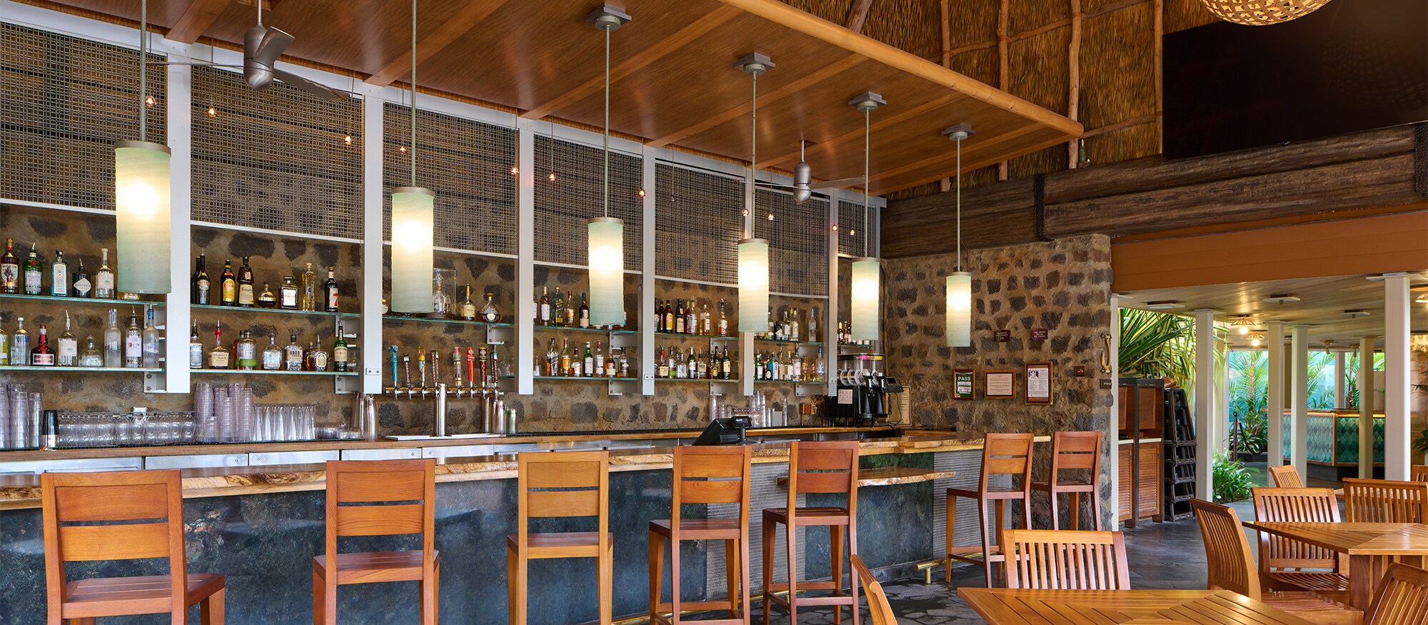 A bar with wooden stools, tables and chairs and a view through plate glass windows of tropical trees