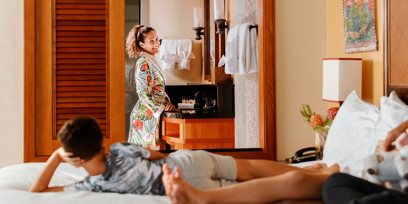 A girl standing by a bathroom vanity while talking to kids who are lounging on a bed