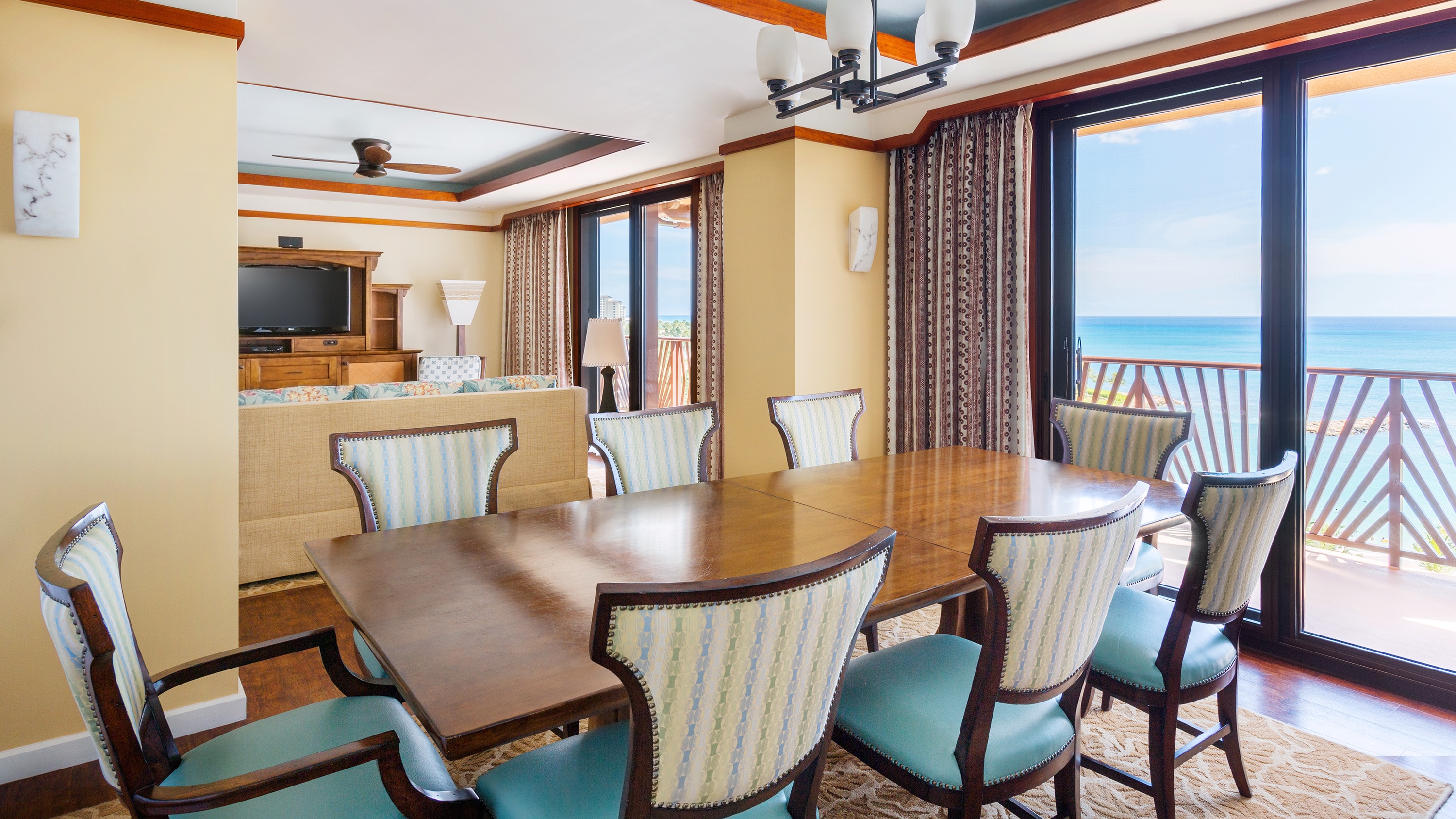 A dining area with a table, 8 chairs and balcony access near  room with a couch, a dresser and a TV