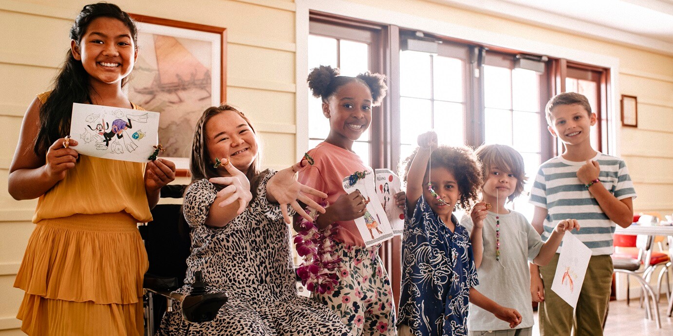 A group of young children smiling at Aunty’s Beach House while showing their paintings.