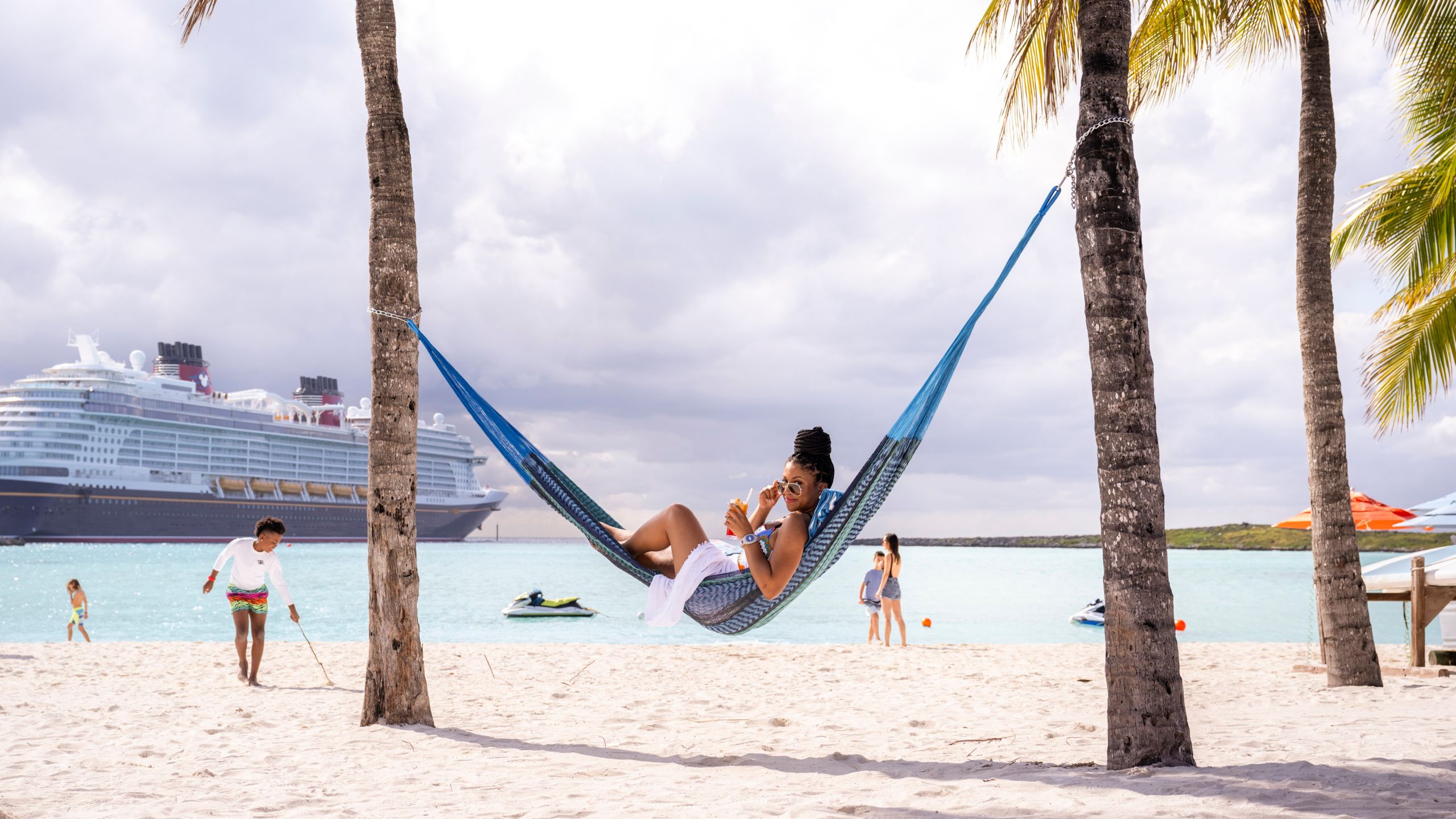 A woman lying in a hammock on the beach with children playing in the ocean in the background
