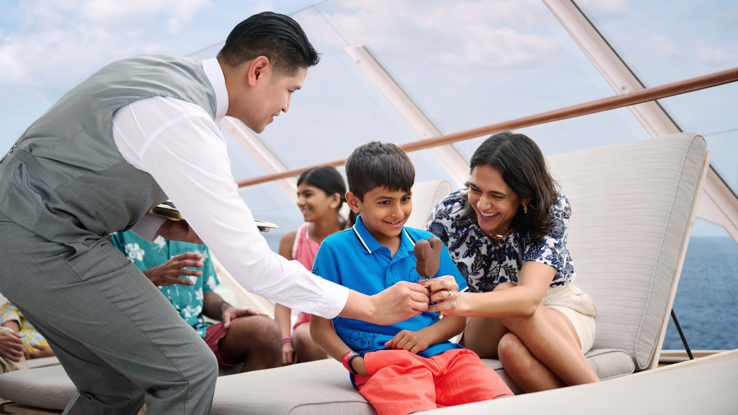 Un Miembro del Elenco entregando un helado con forma de Mickey a una mujer y su hijo en la cubierta superior de un barco de Disney Cruise Line