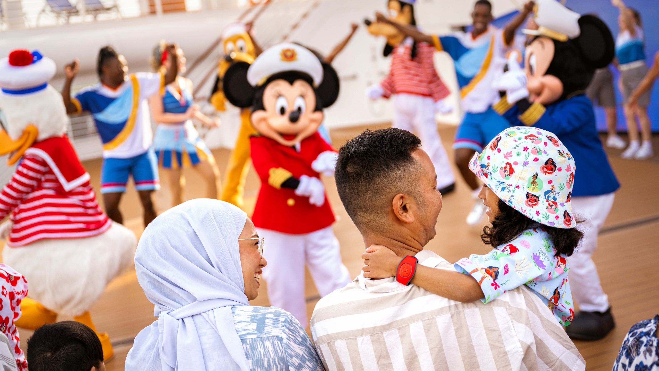 A father holding a child while she smiles at him and her mother, while Disney Characters and Cast Members dance 