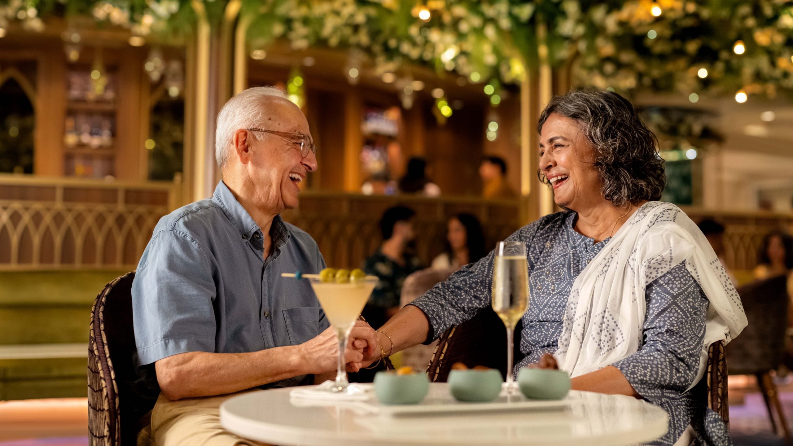 An elderly man and elderly woman holding hands at a dining table with drinks and small dishes of food