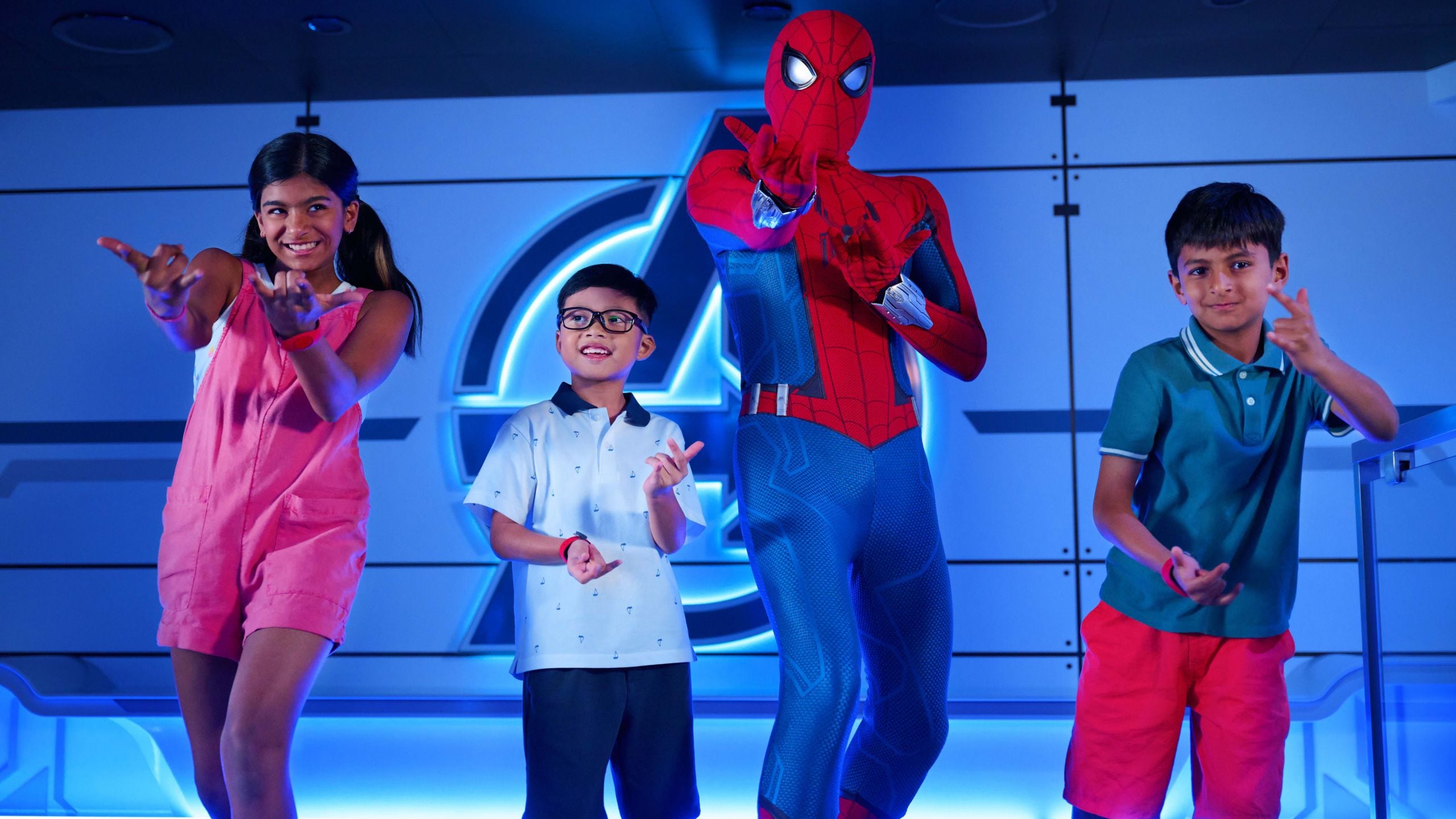 Two boys, a girl and Spider Man posing together in front of an illuminated Avengers logo