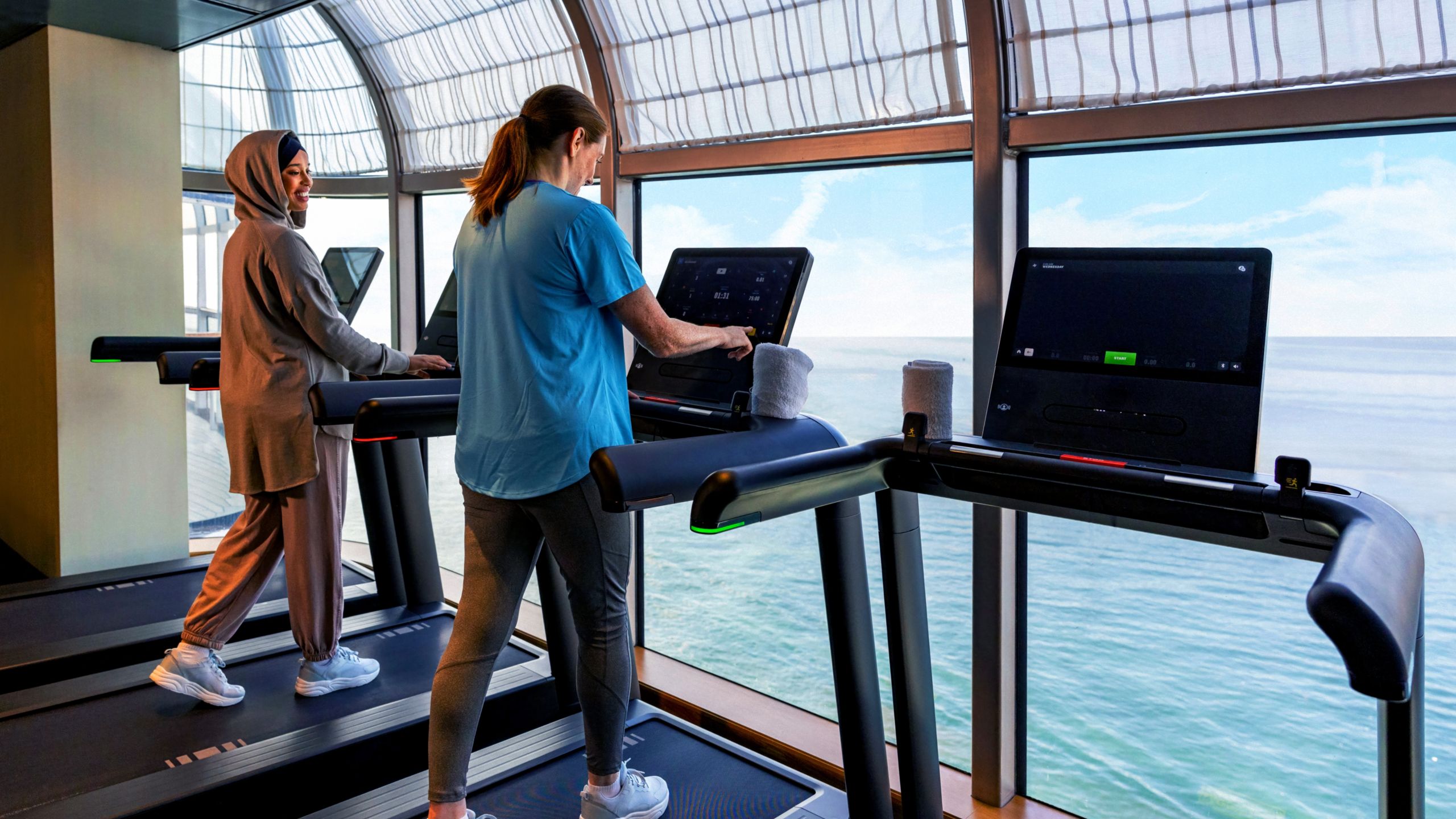 Two women walk on treadmills in a gym, with windows facing out to the sea
