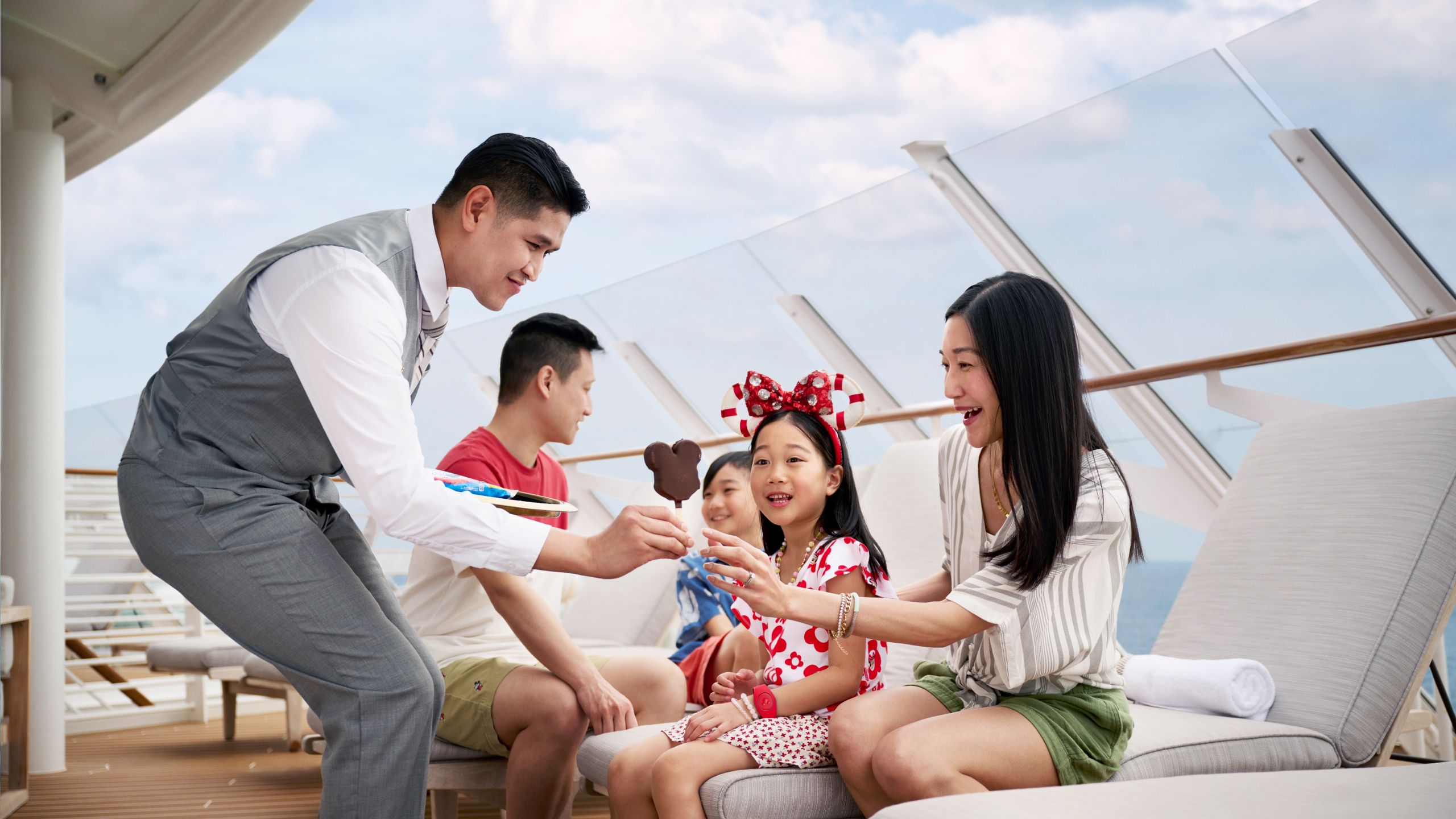 A Cast Member giving a Mickey Mouse shaped ice cream bar to a woman relaxing with her family on the upper deck of a Disney Cruise Line ship