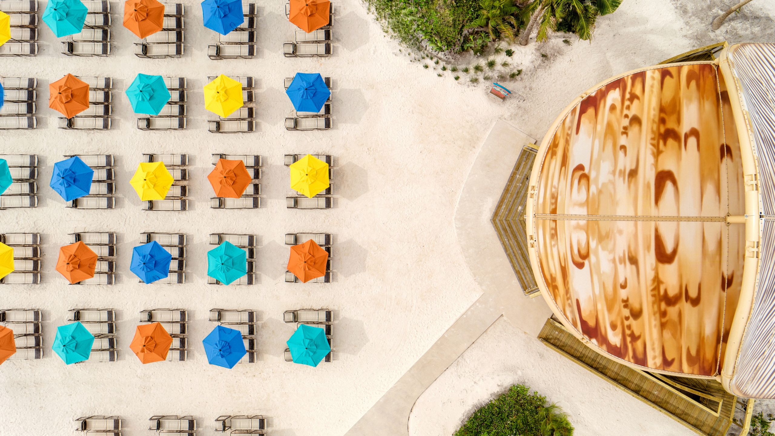 An aerial view of a beach with umbrellas above rows of lounge chairs near a large, circular wooden roofed structure