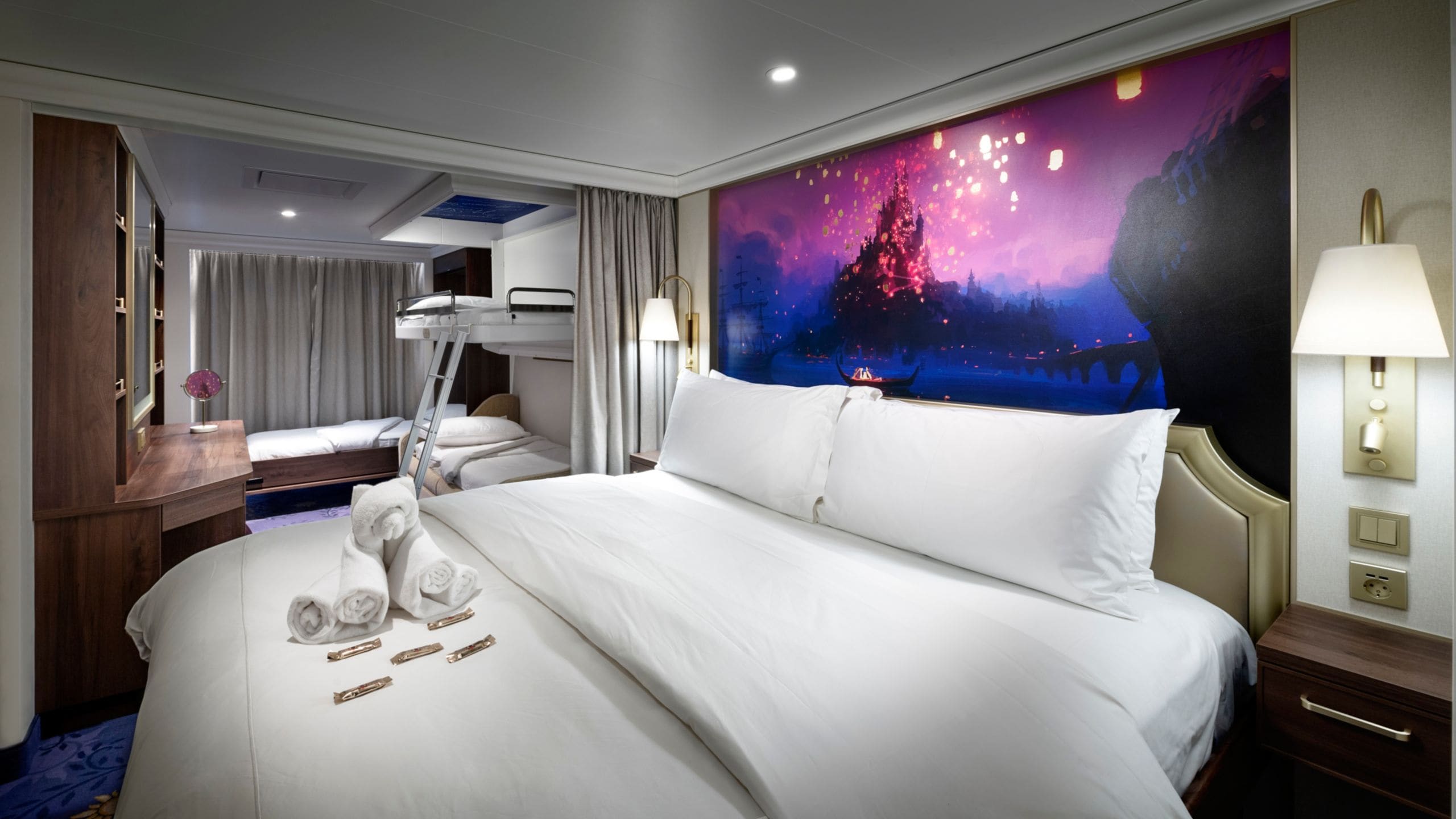 A hotel bedroom with a towel art sculpture on a large bed near bunk beds and a mural of floating lanterns