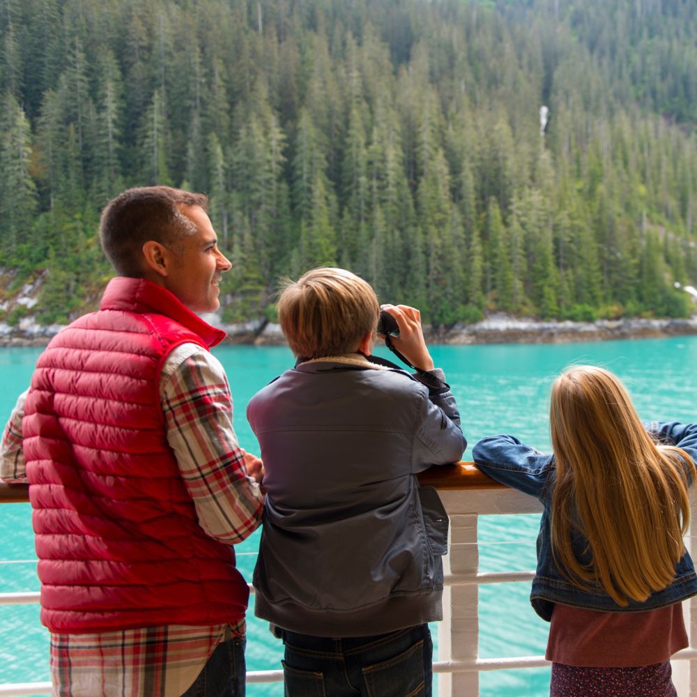 A family of 4 standing at the railing of a Disney Cruise Line ship and admiring a scenic fjord in Alaska