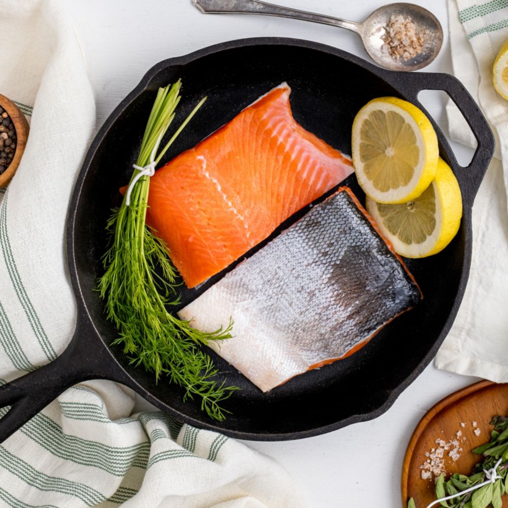 A cast iron skillet containing salmon fillets, lemon slices and a bundle of fresh dill