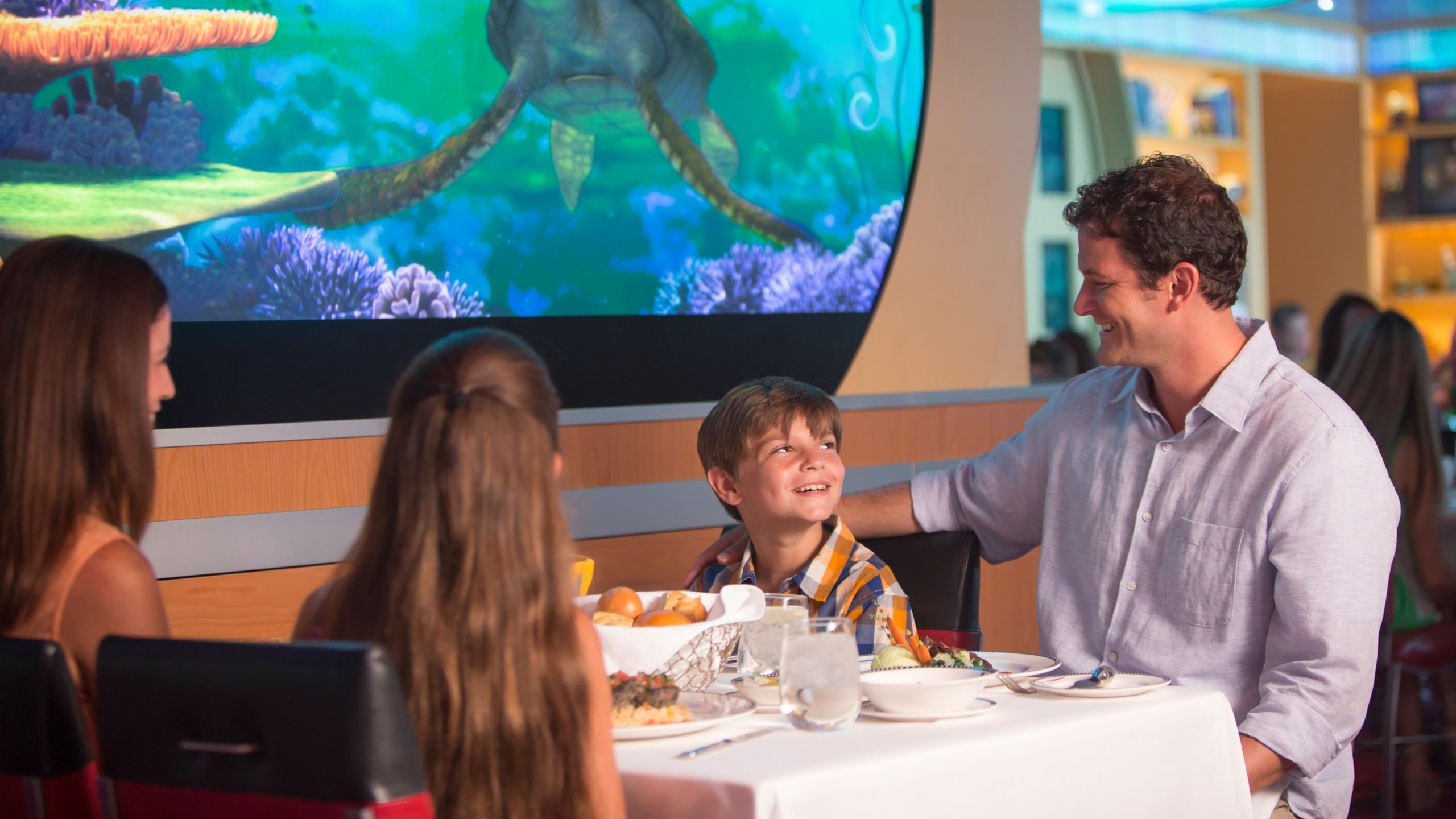 Una familia cenando en Animator’s Palate mientras Crush aparece en una pantalla de video sobre su mesa