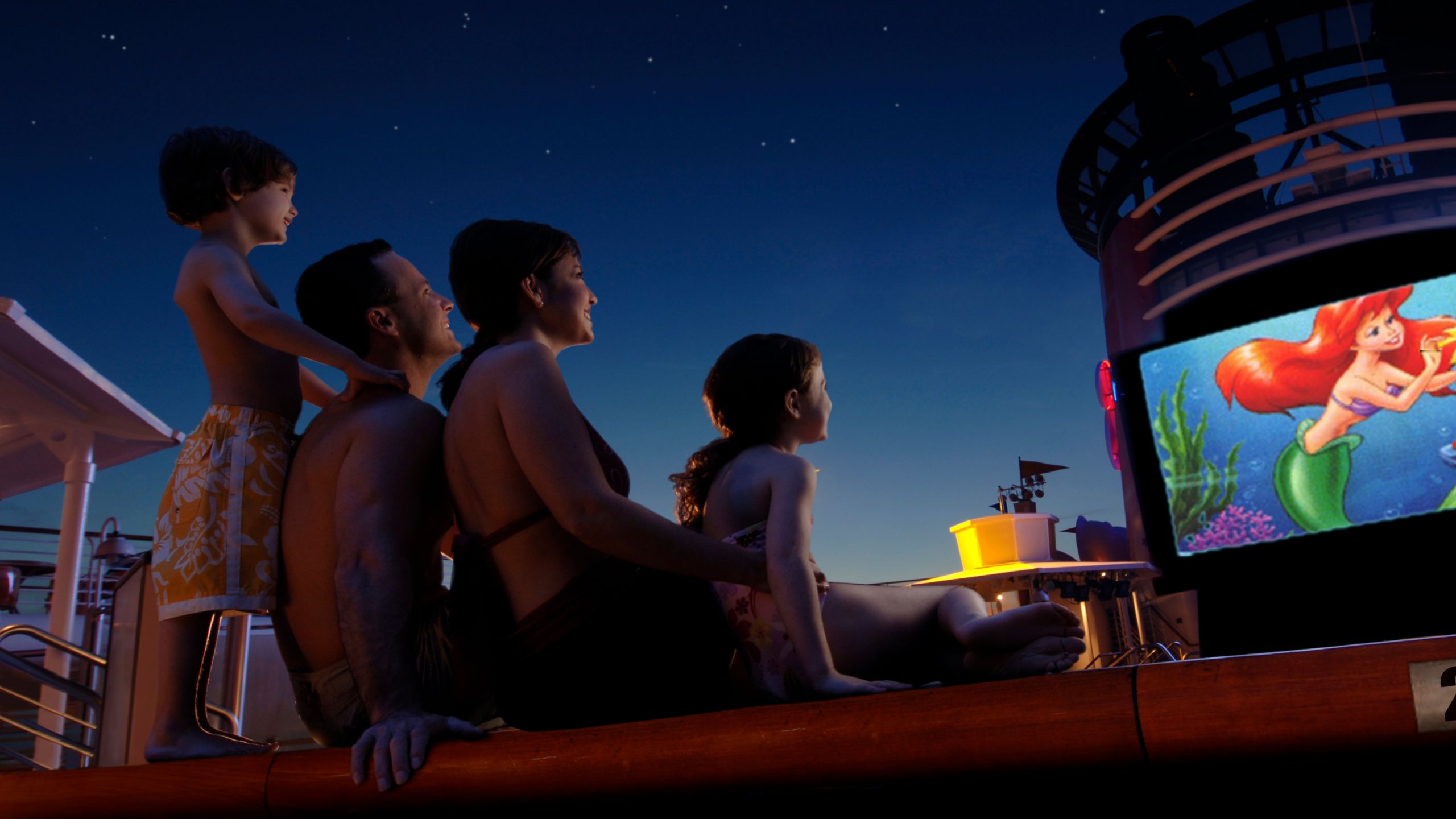 Una familia de Huéspedes viendo La Sirenita en una gran pantalla al aire libre en la cubierta superior de un barco de Disney Cruise Line