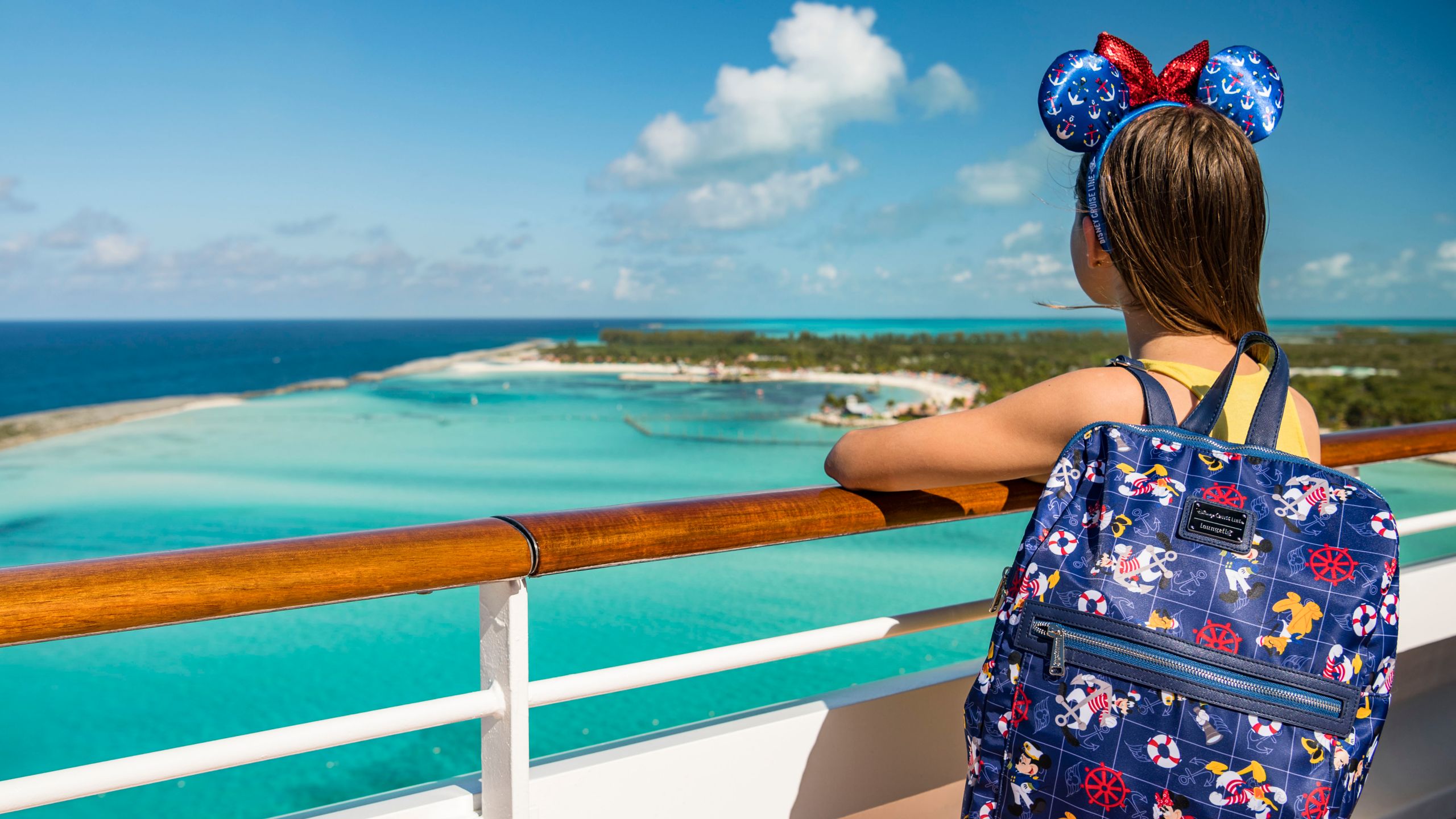 A girl wearing a Minnie Mouse ear headband and a Disney themed backpack looking out at Disney Castaway Cay from a cruise ship railing