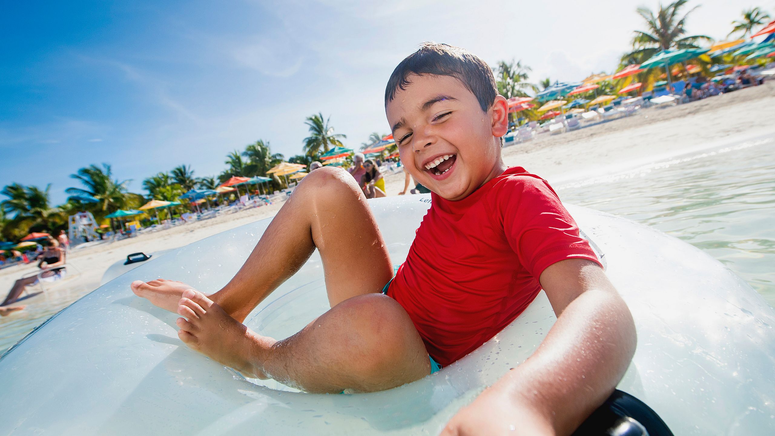 A smiling boy lounging in a water tube near the shore with beach umbrellas and palm trees in the background
