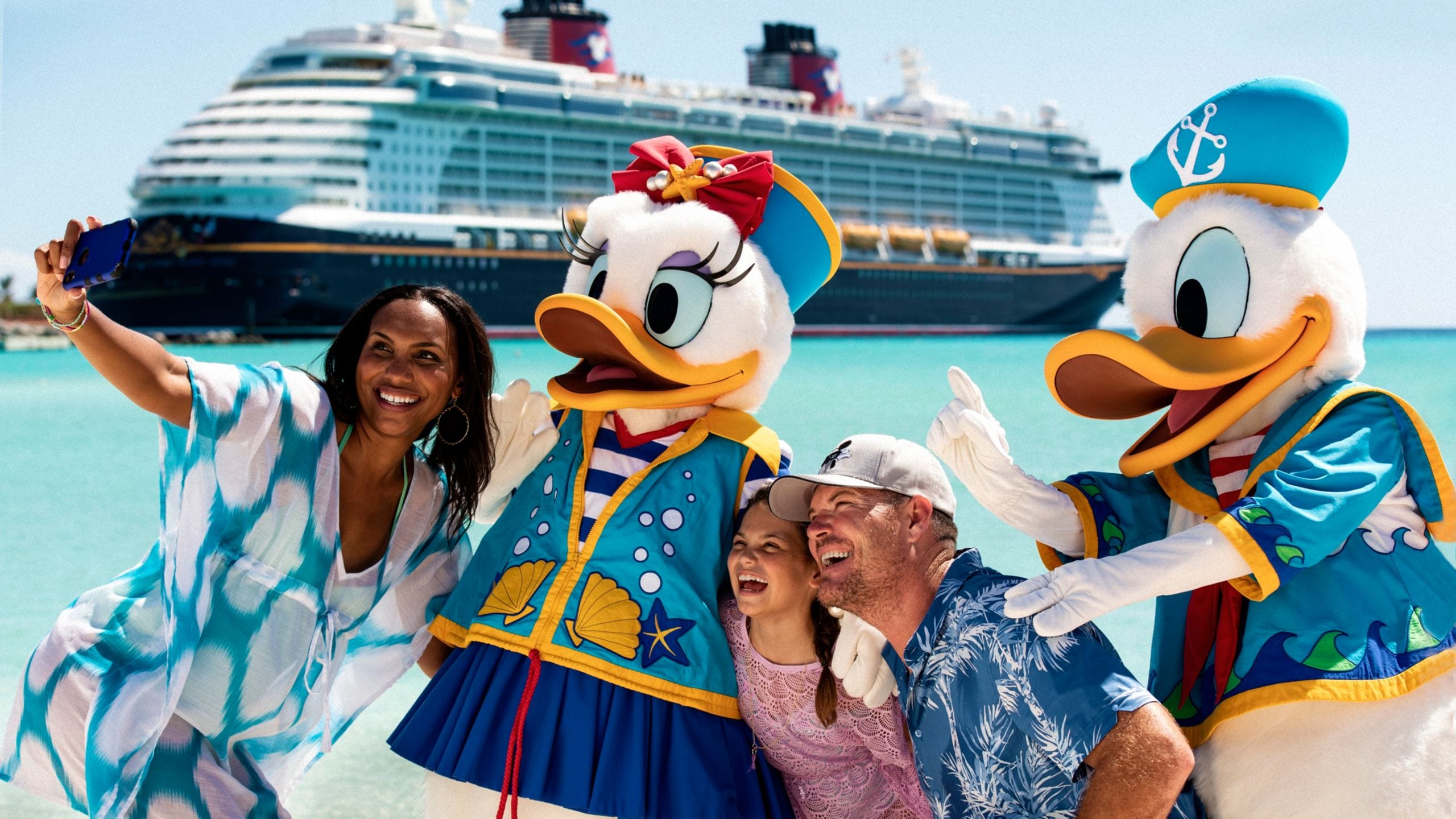 A family posing for a selfie with Donald Duck and Daisy Duck in nautical outfits on the beach at Disney Castaway Cay, with a Disney Cruise Line ship in the background